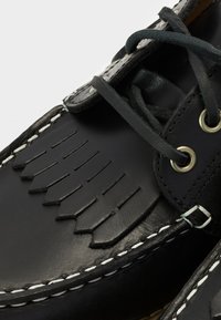 Chaussure en cuir noire avec des détails à franges, des coutures blanches et des lacets noirs passés dans des œillets métalliques.