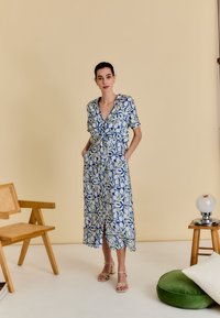 Robe à motifs floraux avec manches courtes, nœud à l'avant et poches. Tons bleu et blanc avec des accents verts. Le modèle porte des sandales à talons beiges.