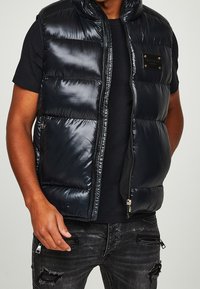 Černá prošívaná vesta s lesklým povrchem, přední zip, vysoký límec a detail loga na hrudi. Kombinováno s černým tričkem a popraskanými džíny.