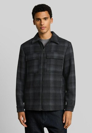 Junger Mann trägt eine dunkelgraue karierte Jacke mit Reißverschluss, zwei Brusttaschen und Kragen, steht vor einem einfarbigen hellgrauen Hintergrund.