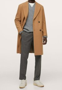 Hellbrauner doppelreihiger Mantel über einem grauen Strickpullover und grauen Hosen, kombiniert mit weißen Sneakers. Glatte Texturen und taillierte Passform.