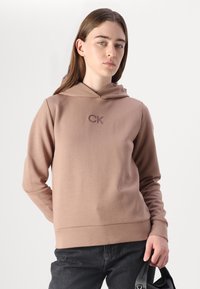 Sudadera con capucha en suave tejido color rosa claro con un logo de CK en un tono más oscuro. Diseño ajustado, mangas largas y ribete acanalado. Combinada con jeans oscuros.