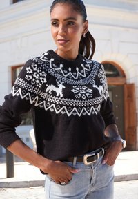 Frau mit dunklem Haar in einem Pferdeschwanz, die einen schwarz-weißen gemusterten Pullover mit Rentieren und Schneeflocken trägt, steht im Freien nahe einem Gebäude.