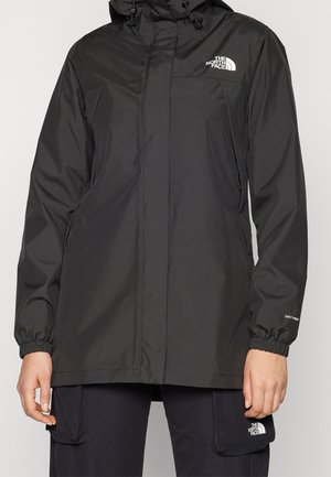 Czarna kurtka przeciwdeszczowa z kapturem, zapinana na zamek błyskawiczny, z elastycznymi mankietami, logo North Face na piersi i lewej kieszeni typu cargo, noszona z pasującymi spodniami.