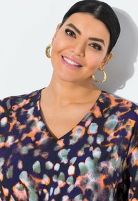 Mehrfarbige Bluse mit V-Ausschnitt, die eine Mischung aus orangefarbenen, blauen und grünen abstrakten Mustern vor einem dunklen Hintergrund zeigt.