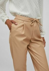 Bež hlače iz umetnega usnja z elastiko v pasu, nagubanim sprednjim delom in stranskima žepoma. Tkanina ima gladko, rahlo sijajno teksturo.