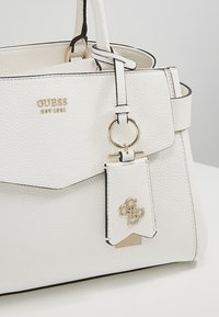 Valkoinen nahkalaukku, jossa on teksturoitu pinta, mustat yksityiskohdat ja kultaiset metalliosat. Varustettu irrotettavalla logotagilla ja renkaalla hihnassa.