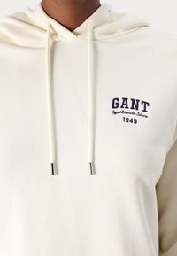 Jasno kremowa bluza z kapturem z sznurkiem przy dekolcie, z granatowym haftowanym napisem "GANT Sportswear Since 1949" na klatce piersiowej. Miękka faktura.
