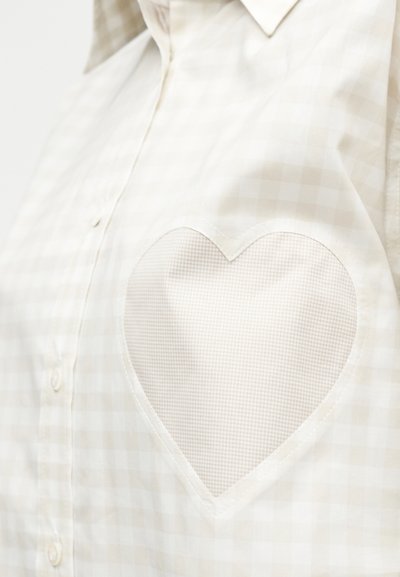 Πουκάμισο σε μπεζ και λευκό καρό με κουμπιά, με μια μεγάλη τσέπη σε σχήμα καρδιάς στο στήθος, σε ανοιχτότερο καρό σχέδιο.