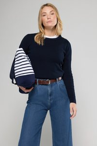 Helleblauer, gerippter Pullover, hochtaillierte blaue Jeans, brauner Gürtel, hält eine gestreifte Tasche in Marineblau und Weiß, mit langen blonden Haaren.