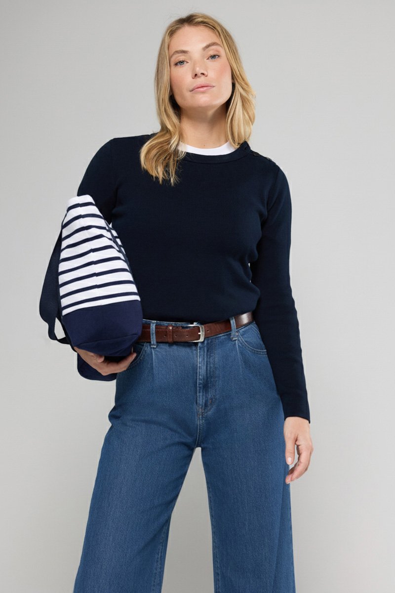 Helleblauer, gerippter Pullover, hochtaillierte blaue Jeans, brauner Gürtel, hält eine gestreifte Tasche in Marineblau und Weiß, mit langen blonden Haaren.