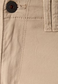 Beežid puuvillased püksid, millel on sile tekstuur, tume nööp ja peen õmblused taskutel ning vöökohal.