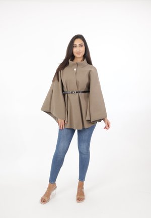 Brun kappa-liknande ytterplagg med hög krage, med en enkel knappslutning och ett svart bälte i midjan, matchad med blå jeans.
