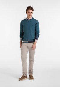 Maglione teal con polsini a righe a contrasto sopra una camicia con colletto, abbinato a pantaloni beige chiaro e mocassini in suede marrone. Design semplice.