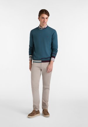 Maglione teal con polsini a righe a contrasto sopra una camicia con colletto, abbinato a pantaloni beige chiaro e mocassini in suede marrone. Design semplice.