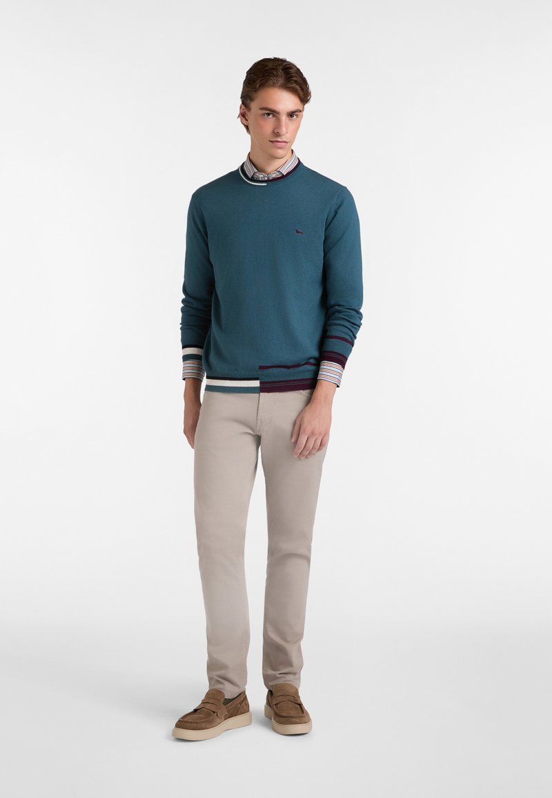 Maglione teal con polsini a righe a contrasto sopra una camicia con colletto, abbinato a pantaloni beige chiaro e mocassini in suede marrone. Design semplice.