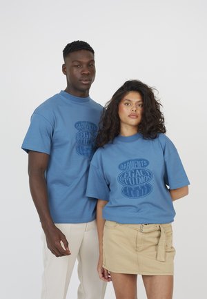 Man en vrouw staan zij aan zij en dragen bijpassende blauwe "Legacy Studios" T-shirts; de man draagt lichte broek, de vrouw een beige rok.