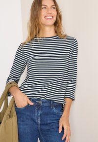 Maglietta a righe blu navy e bianca con scollo rotondo e maniche a tre quarti, abbinata a jeans blu e un piumino verde.