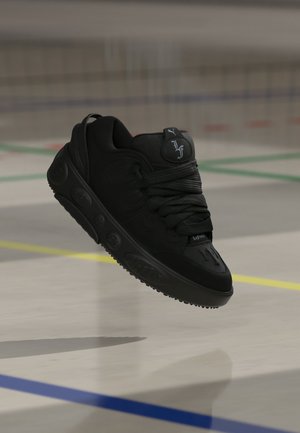 Schwarzer Sportsneaker mit dicker Sohle und detaillierten Logos, schwebend in der Luft über einem Hallensportplatz mit farbigen Linien.