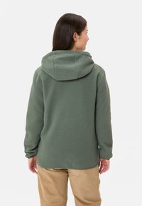 Felpa verde in pile con una finitura testurizzata, dotata di un ampio cappuccio e maniche lunghe. La parte posteriore è leggermente più lunga con un orlo curvo.