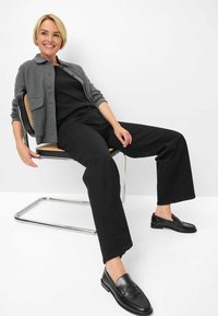 Cardigan grigio con bottoni sopra una maglietta nera, abbinato a pantaloni ampi neri e mocassini neri, seduto su una sedia con struttura metallica.