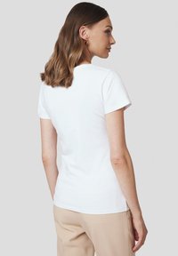 Biała koszulka z krótkim rękawem, okrągłym dekoltem, dopasowany krój i gładki materiał bawełniany, pokazana od tyłu na szarym tle.