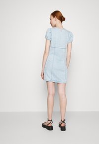 Vestido de mezclilla azul claro con mangas cortas abullonadas, diseño ajustado y espalda estructurada. Llevado con sandalias de plataforma negras. Textura suave.
