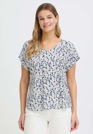 Femme souriante aux longs cheveux portant des boucles d'oreilles créoles, un chemisier à manches courtes à motif floral en V et un pantalon blanc, debout devant un fond uni.