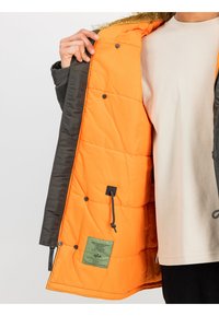 Chaqueta acolchada con un exterior gris oscuro y un forro naranja vibrante. Cuenta con una capucha forrada de pelo, botones a presión y una etiqueta verde en el forro interior.