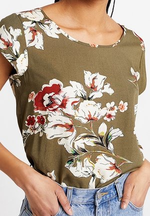 Oliwkowa bluzka z krótkim rękawem z dużym, białym i czerwonym kwiatowym wzorem, noszona wpuszczona w jasnoniebieskie dżinsy.