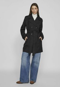 Juodas trench coat'as su dvigubo užsegimo dizainu, dideliais mygtukais ir diržu. Komponuojamas su plačiais mėlynais džinsais ir rudomis smailiomis bateliais.