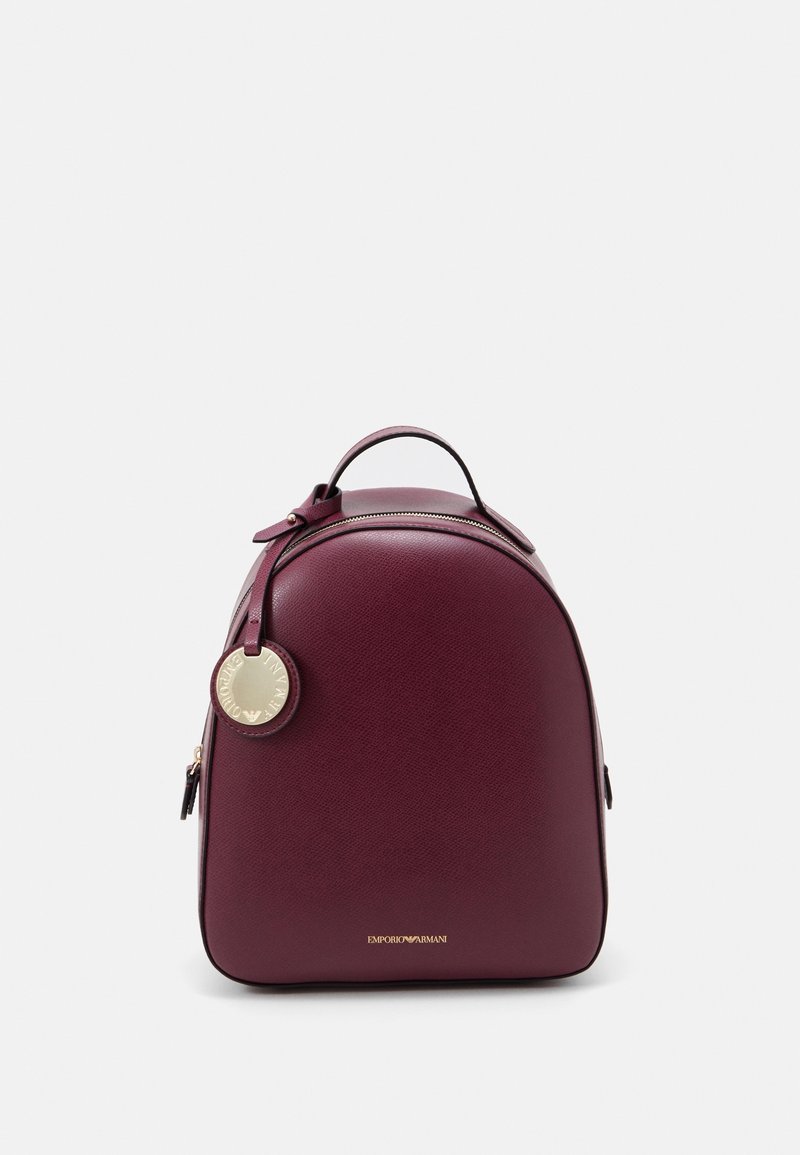 Ryggsäck i bordeauxläder med rundad form, strukturerad yta, topphandtag, guldfärgade beslag och en cirkulär logotypbricka. Dragkedja upptill.