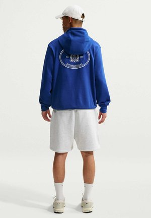 Mann trägt einen blauen Hoodie mit weißem Logo, hellgraue Shorts, weiße Kappe, Socken und beige Sneakers, steht mit dem Rücken zum Betrachter auf weißem Hintergrund.