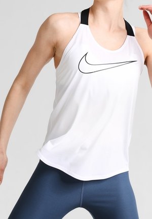 Sportowiec w białej sportowej bluzce bez rękawów marki Nike z czarnymi ramiączkami i granatowych legginsach w dynamicznej pozie na jasnym tle.