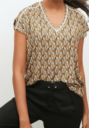 Vrouw draagt een bruin, patroonrijk T-shirt met V-hals en korte mouwen, ingestopt in een hoge taille zwarte broek met een ronde zwarte riemgesp.