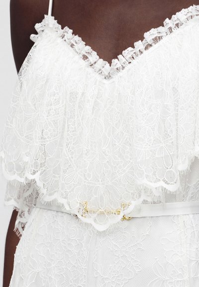 Άτομο με σκουρόχρωμη επιδερμίδα που φοράει λευκό δαντελένιο φόρεμα με ανθικά μοτίβα και σούρες στο V-λαιμόκοψη, τονισμένο με λεπτή λευκή ζώνη με χρυσή αγκράφα.