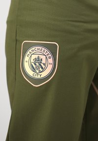 Olivgrüne Hose mit einem navyfarbenen Manchester City-Logopatch mit pinkem Saum auf der Vorderseite der Tasche. Glatte Stoffstruktur.