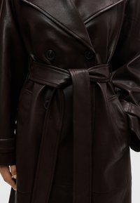 Καφέ σκούρο δερμάτινο παλτό τύπου trench με φαρδιές πέτρες γιακά, διπλό κούμπωμα με μαύρα κουμπιά, πλαϊνές τσέπες και δέσιμο στη μέση με ζώνη.