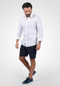 Chemise blanche à manches longues à boutons, associée à un short bleu marine et des baskets en daim bleu. Le modèle se tient devant un fond neutre, les yeux baissés.