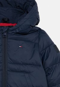 Mornarško modra kapuca s podlogo in sprednjim zadrgo, žepom ter majhnim rdečim, belim in modrim logotipom na levem prsnem delu.