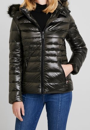 Vrouw die een zwarte glanzende gewatteerde jas met capuchon en bontafwerking draagt, gecombineerd met een blauwe spijkerbroek en een zwarte coltrui.