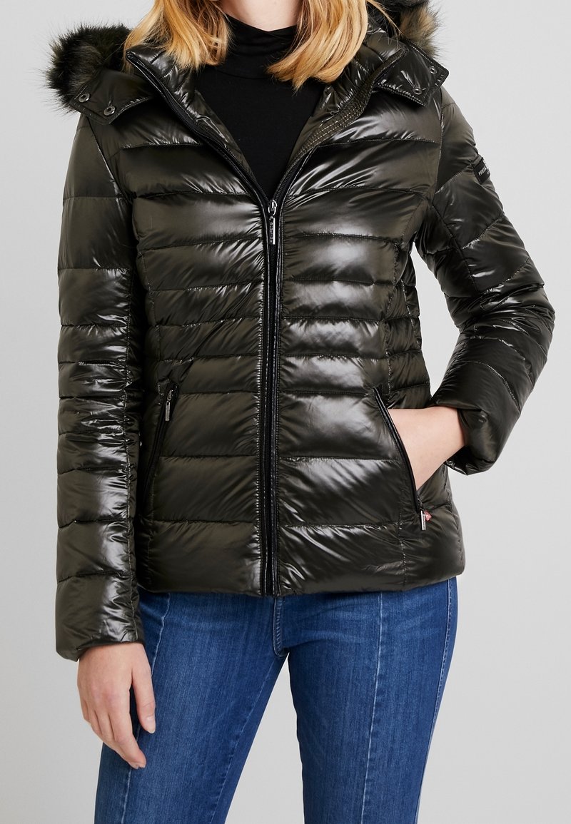 Femme portant une veste matelassée noire brillante avec capuche et bordure en fourrure, associée à un jean bleu et un haut à col roulé noir.