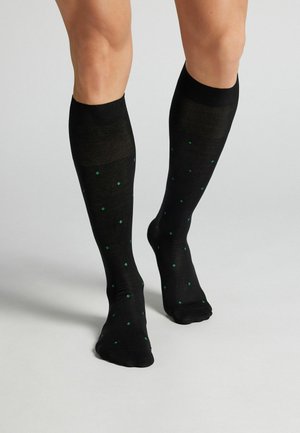 Ben iført sorte knæstrømper med små grønne diamantmønstre, stående på en lysegrå overflade.