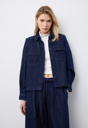 Junge Frau steht vor einer weißen Wand und trägt eine dunkelblaue Jeansjacke sowie passende weitere Hosen mit weitem Bein, darunter ein weißes, kurzes Oberteil.