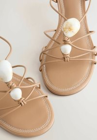 Sandales plates beiges avec de fines lanières et de grosses perles blanches effet marbre sur le dessus, comportant des coutures blanches le long des bords de la semelle.