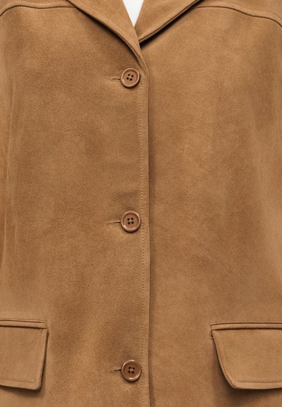 Κοντινό πλάνο ενός ταμπά σουέντ παλτό με τρία καφέ κουμπιά στη μέση και δύο πλαϊνές τσέπες με καπάκι κοντά στο κάτω μέρος.