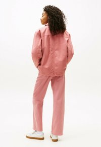 Giacca oversize rosa e pantaloni abbinati mostrati di spalle, realizzati in un tessuto resistente con una trama liscia. Scarpe da ginnastica bianche completano il look.