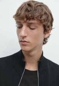 Giacca nera con zip e collo alto, con leggera costina, indossata sopra una maglietta scura; il modello ha i capelli castani mossi. Design minimalista.