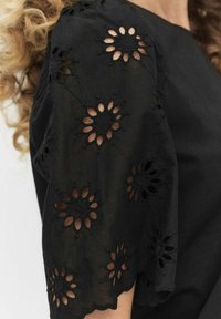 Czarna bluzka z krótkimi rękawami, ozdobiona haftowanymi wzorami kwiatowymi oraz wycięciami. Gładki materiał o luźnym kroju i falbaniastym dole.