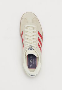Adidași din suede bej cu accente roșii, șireturi albe și talpă din cauciuc. Prezintă un logo pe limba pantofului și căptușeală din textil.
